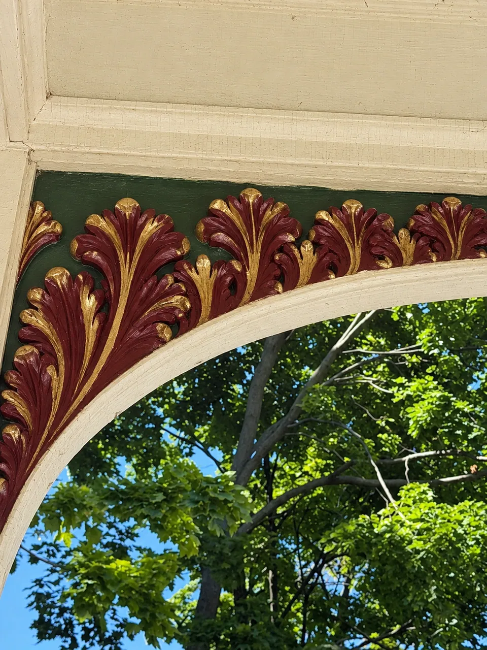 A rendering of the porch arch painted: burgundy acanthus leaves edged in gold on a deep green ground under cream trim, with trees and sky beyond.