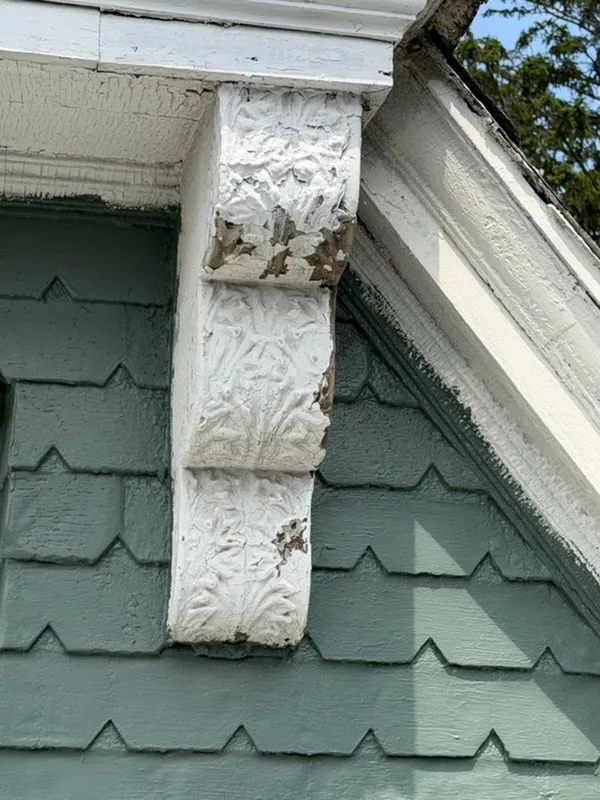 A three-tier carved bracket under the gable, thick with peeling white paint, against green shingles.