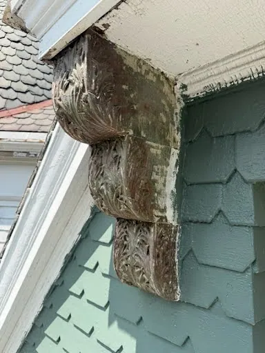 A carved bracket stripped to bare wood, seen from below against the green shingles.